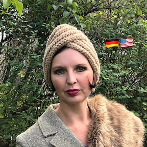 Könnte beinhalten: Eine Frau trägt einen beigefarbenen Häkel-Turban, einen braunen Pelzkragen und eine Tweedjacke. Sie steht vor einem grünen, belaubten Hintergrund.