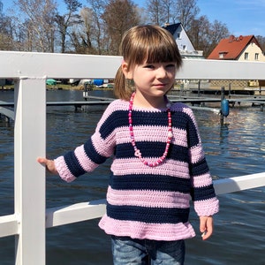 May include: A young girl wearing a pink and navy blue striped crocheted sweater with a pink beaded necklace. She is standing on a white wooden dock with a body of water behind her.