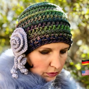 以下が含まれることがあります： 緑、紫、青のクロシェ編みビーニーに、合わせたクロシェ編みの花の装飾が施されています。ビーニーは、黒髪で茶色の目の人の頭にかぶっています。
