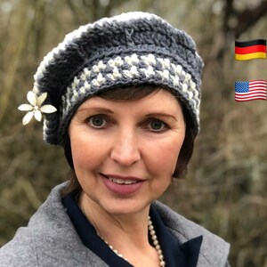 May include: A woman wearing a gray and white crocheted beret with a white flower accent. The beret has a checkered pattern. The woman is wearing a gray jacket and a pearl necklace. The background is blurred and shows green foliage. The image also includes a German flag and an American flag.