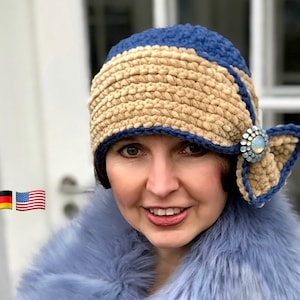 May include: A woman wearing a blue and beige crocheted cloche hat with a blue and white jeweled pin. The hat has a wide brim and a bow on the side. The woman is wearing a blue faux fur collar.
