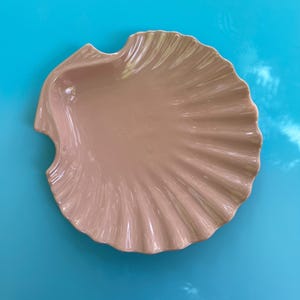 May include: A light beige, seashell-shaped ceramic dish. The dish has a scalloped edge and a ridged interior, creating a textured surface. The dish is set against a bright blue background.
