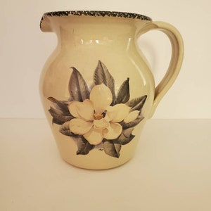 May include: Cream-colored ceramic pitcher with a large magnolia flower design. The pitcher has a curved handle and a spout. The rim is decorated with a dark, speckled pattern. The magnolia flower is white and yellow with gray-green leaves.