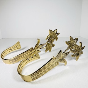 May include: A pair of polished brass curtain tiebacks featuring a U-shaped hook design with a ribbed texture. Each tieback is adorned with a decorative floral motif at the end, showcasing detailed petals and leaves in a metallic gold finish.