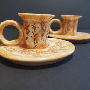 Puede incluir: Dos portavelas de cerámica con un diseño floral marrón y blanco. Los portavelas tienen una base de platillo y una parte superior en forma de taza.