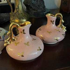 May include: Two small pink ceramic vases with gold leaf accents. The vases have a rounded body and a small handle. The vases are decorated with a pattern of gold leaves.