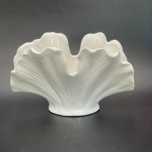 May include: A white ceramic decorative bowl with a scalloped, fan-like design. The bowl has a wide, open top and a textured, ridged exterior, resembling a stylized seashell. The bowl is set against a dark background.