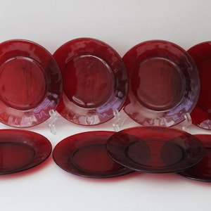 May include: A set of eight ruby red glass plates. The plates are round with a slightly raised rim. The plates are displayed on clear plastic plate stands. The plates are a deep, rich red color.