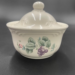 May include: A white ceramic sugar bowl with a lid. The bowl features a floral design with green leaves and purple grapes. The lid has a small knob on top.
