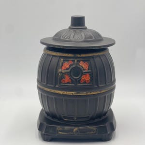 May include: A black decorative container with a lid, resembling a small barrel. The container has a gold trim and a decorative floral design in red and orange. The base is rectangular and the lid has a small handle.