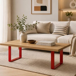 May include: A rectangular coffee table with a light wood grain top and two bright red U-shaped metal legs. The table surface holds a white ceramic vase with green foliage, a small dark brown bowl, and two stacked books with white covers.