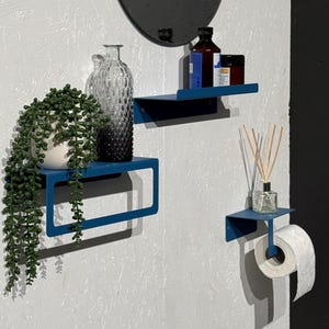 Puede incluir: Una escena de baño moderno con un espejo circular gris oscuro y tres estantes de metal azul. Los estantes contienen una planta en maceta, una botella decorativa y artículos de tocador. Un rollo de papel higiénico está en el estante inferior.