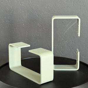 May include: Two light green metal table legs with a U-shaped design. Each leg has a flat top with pre-drilled holes for mounting. The legs are displayed on a black table against a textured gray background.