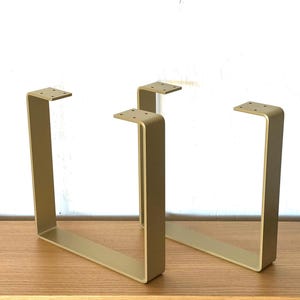 May include: Gold-colored, U-shaped metal table legs. Each leg has a rectangular top with pre-drilled holes for mounting. The legs are made of a sturdy metal and have a modern, minimalist design. The legs are approximately 16 inches tall.