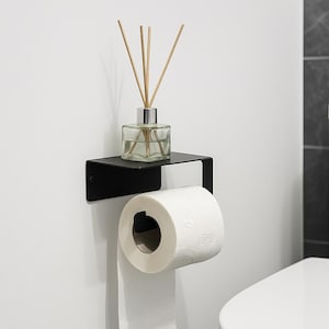 Op de afbeelding: Een zwarte metalen toiletpapierhouder met een plank, met een rol wit toiletpapier. Een glazen diffuserfles met riet staat op de plank. De houder is gemonteerd op een witte muur, naast een wit toilet.