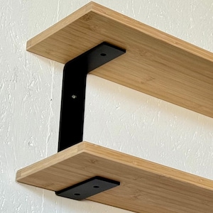 May include: Two-tier wooden shelf with black metal brackets. The shelves are made of light-colored wood and are mounted on a white and black wall. The brackets are rectangular and provide support for the shelves.