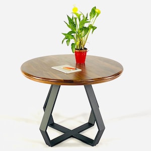 Può includere: Un tavolino rotondo in legno con un piano marrone scuro e una base geometrica in metallo nero. Un piccolo vaso rosso con una pianta verde è posto sopra, insieme a un blocco note e una matita. Il tavolo ha un diametro di circa 61 cm.