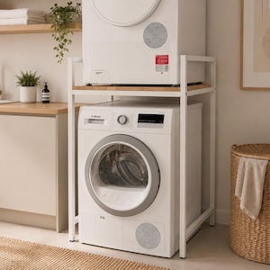 Op de afbeelding: Een witte wasmachine en droger gestapeld op een wit metalen frame. De droger staat bovenop de wasmachine. Een geweven mand met deksel en handdoek staat rechts. De wasruimte heeft een neutraal kleurenpalet.