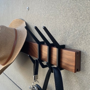 May include: A wooden wall mounted coat rack with three black metal hooks. A straw hat, a black leather bag, and a set of keys are hanging on the hooks.