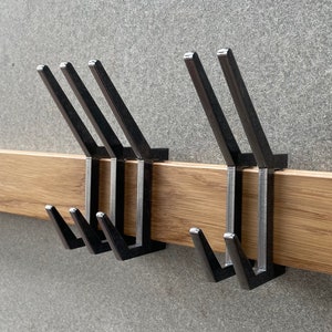 Könnte beinhalten: Ein Wand-Kleiderhaken aus Holz mit fünf schwarzen Metallhaken. Die Haken sind versetzt angeordnet, mit zwei Sätzen von zwei Haken und einem einzelnen Haken.