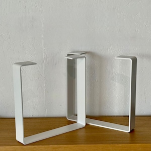 Könnte beinhalten: Drei weiße Metall-Tischbeine in modernem, minimalistischem Design. Jedes Bein hat eine rechteckige Form mit flacher Oberseite und einem rechtwinkligen Knick an der Basis. Auf einer Holzoberfläche vor einer weißen Wand präsentiert.