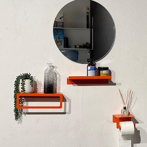 Peut inclure: Aménagement de salle de bain moderne avec un miroir rond et trois étagères flottantes orange vif. Les étagères accueillent une petite plante en pot, une bouteille décorative et des articles de toilette. Un rouleau de papier toilette est suspendu.