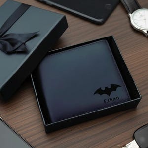 Puede incluir: Una cartera de cuero azul marino con el logotipo de Batman y el nombre "Ethan" se muestra dentro de una caja de regalo negra. Un lazo negro adorna la tapa de la caja. Otros elementos incluyen un reloj, llaves y dos teléfonos inteligentes.