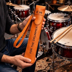 Puede incluir: Un estuche para baquetas de cuero color canela con cierre de solapa y las iniciales E.M. grabadas en negro. El cuerpo vertical del estuche muestra el texto FOR THE DRUMMER en letras negras e incluye una correa de muñeca de cuero a juego.