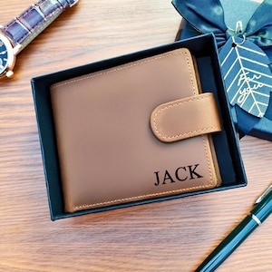 Könnte beinhalten: Ein braunes Lederportemonnaie mit dem eingeprägten Namen "JACK" auf der Vorderseite. Die Geldbörse befindet sich in einer dunkelblauen Geschenkbox mit Schleife. Eine Uhr und ein Stift sind ebenfalls zu sehen.