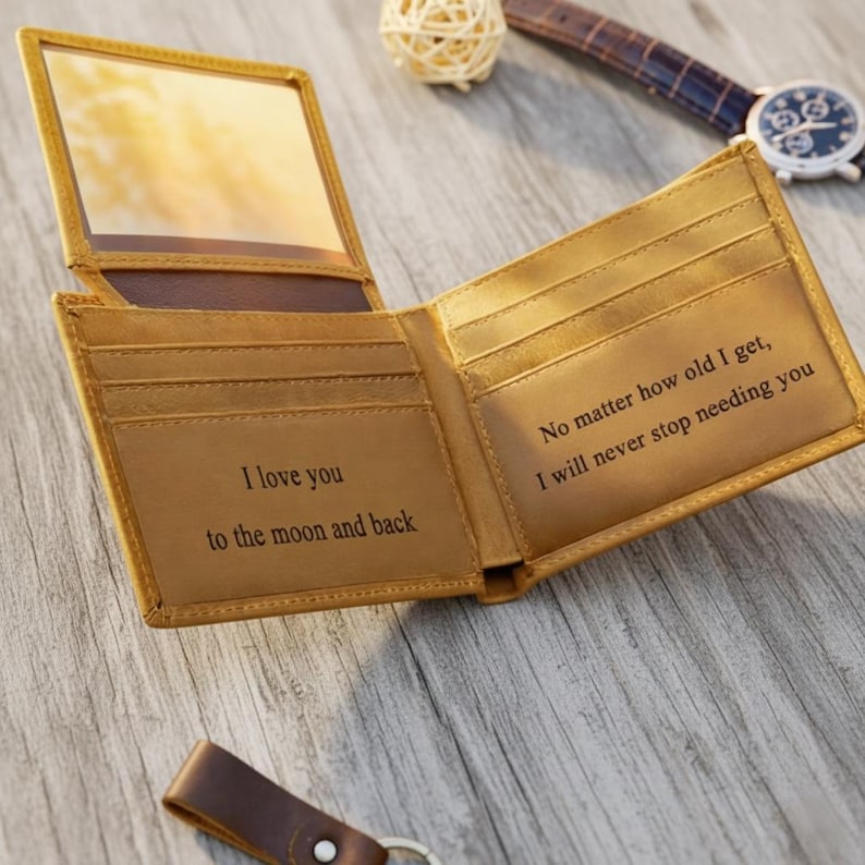 May include: Open, tan leather wallet with a mirror, featuring the engraved phrases "I love you to the moon and back" and "No matter how old I get, I will never stop needing you."