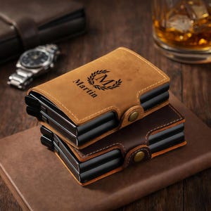 Puede incluir: Dos carteras de cuero, una marrón claro y otra marrón oscuro, apiladas. La cartera marrón claro tiene el nombre "Martin" y un diseño de monograma. Ambas carteras tienen un interior negro y un cierre metálico. Un reloj y un vaso de líquido ámbar en el fondo.