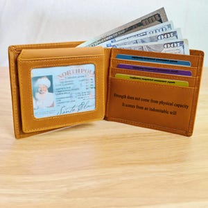Może przedstawiać: Otwarty, jasnobrązowy skórzany portfel z prawem jazdy Świętego Mikołaja. Portfel zawiera kilka banknotów i kart kredytowych. Wnętrze portfela zawiera cytat: "Siła nie pochodzi z fizycznej sprawności."