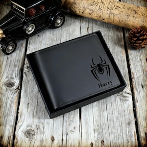 Puede incluir: Cartera de cuero negro con un diseño de araña y el nombre "Harry" grabado. La cartera está en una caja negra, sobre una superficie de madera rústica. También se ve un coche de juguete y una piña.