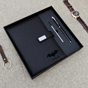 May include: A black leather journal with a Batman logo and the name "Ethan" is presented in a black gift box, alongside a pen and stylus. A brown leather watch and keyring are also visible.