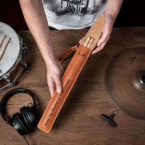 Puede incluir: Un estuche de cuero color canela para baquetas con el texto grabado E.M y Keep the Rhythm of Your Dreams. El estuche contiene dos baquetas de madera e incluye una correa de cuero a juego para llevarlo, presentado en una vista clara.