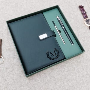 Puede incluir: Un diario de cuero verde oscuro con un cierre plateado y la letra "M" dentro de una corona de laurel. El diario está en una caja verde oscuro con un bolígrafo y un lápiz óptico. Un reloj es visible en el fondo.