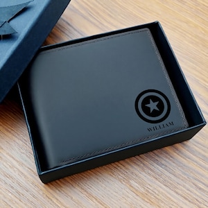May include: A dark gray leather wallet with a black Captain America shield design and the name "WILLIAM" printed below it. The wallet is presented inside a dark blue gift box, set against a wooden surface.