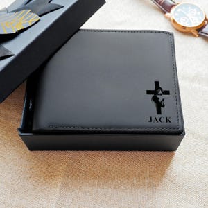 Könnte beinhalten: Schwarzes Lederportemonnaie mit Kreuz- und Gesichtsdesign, präsentiert in einer schwarzen Geschenkbox. Das Portemonnaie ist mit dem Namen "JACK" unter dem Kreuz personalisiert. Im Hintergrund ist eine Armbanduhr mit braunem Armband zu sehen.