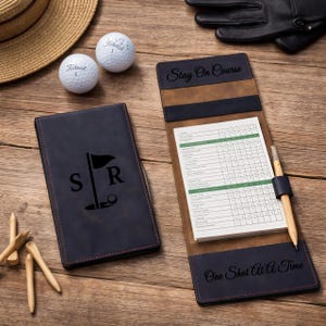 Könnte beinhalten: Ein marineblauer Golf-Scorekartenhalter aus Leder mit den Initialen "S R" und einer Golf-Flaggen-Grafik. Der Halter ist geöffnet und zeigt eine Scorekarte und einen Bleistift. Zwei Golfbälle und Golf-Tees sind ebenfalls sichtbar.