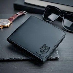 Puede incluir: Cartera de cuero negro con diseño de lobo y el nombre "Olivia" grabado en la parte delantera. La cartera cerrada está sobre una superficie oscura, junto a un reloj y gafas de sol.