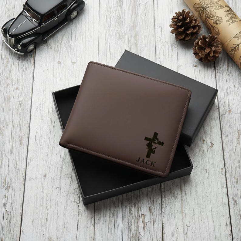 May include: A brown leather wallet with a cross design and the name "JACK" is displayed open in a black box. The wallet is on a light wood surface, with a black toy car and pine cones in the background.