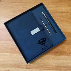 May include: A navy blue notebook and pen set in a gift box. The notebook features a Superman logo and the name "Matthew". The set includes a pen and a refill. The box is navy blue with a silver clasp.