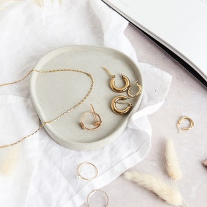 May include: A collection of gold jewelry, including hoop earrings, a delicate chain necklace, and rings, arranged on a gray, circular tray. The jewelry is displayed on a white cloth, with a magazine and a tube of cream in the background.