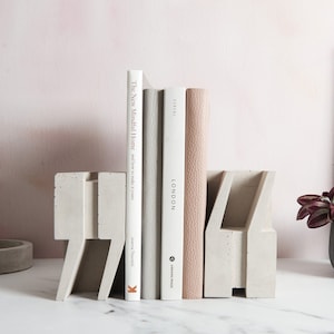 May include: Concrete bookends in the shape of the number seven, holding up three books. The books are titled "The New Minimal Home", "London", and "Edition". A small potted plant with red and green leaves sits on a white surface next to the bookends.