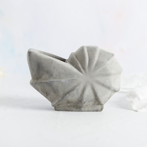 Könnte beinhalten: Ein grauer Betontopf in Form einer Muschel. Der Topf hat eine glatte, strukturierte Oberfläche und eine leicht geöffnete Oberseite.