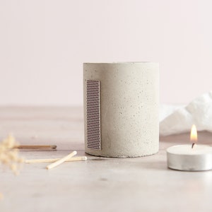 May include: A grey concrete candle holder with a small, pink, gridded match strike surface on the side. A small, white tea light candle is lit and sitting on a white surface.