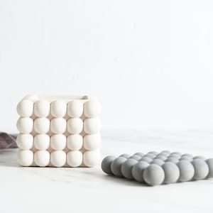 Puede incluir: Dos macetas de cerámica blanca con un diseño texturizado con forma de burbujas. La maceta de la izquierda es más alta y de forma cuadrada. La maceta de la derecha es más baja y de forma rectangular.