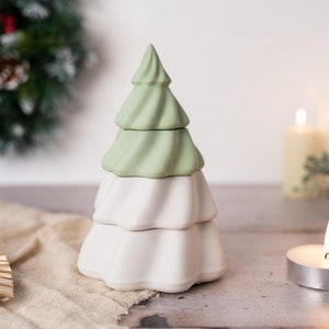 以下が含まれることがあります： 白い土台と3つの緑色の段があるセラミック製のクリスマスツリーのオーナメント。オーナメントはベージュの布の表面の上に置かれています