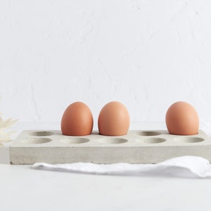 Puede incluir: Tres huevos marrones en un soporte para huevos de hormig&oacute;n gris con 12 ranuras individuales.