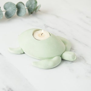 Puede incluir: Un portavelas de cerámica verde claro en forma de tortuga con una pequeña vela blanca en su interior. La tortuga está sentada sobre una superficie de mármol blanco.